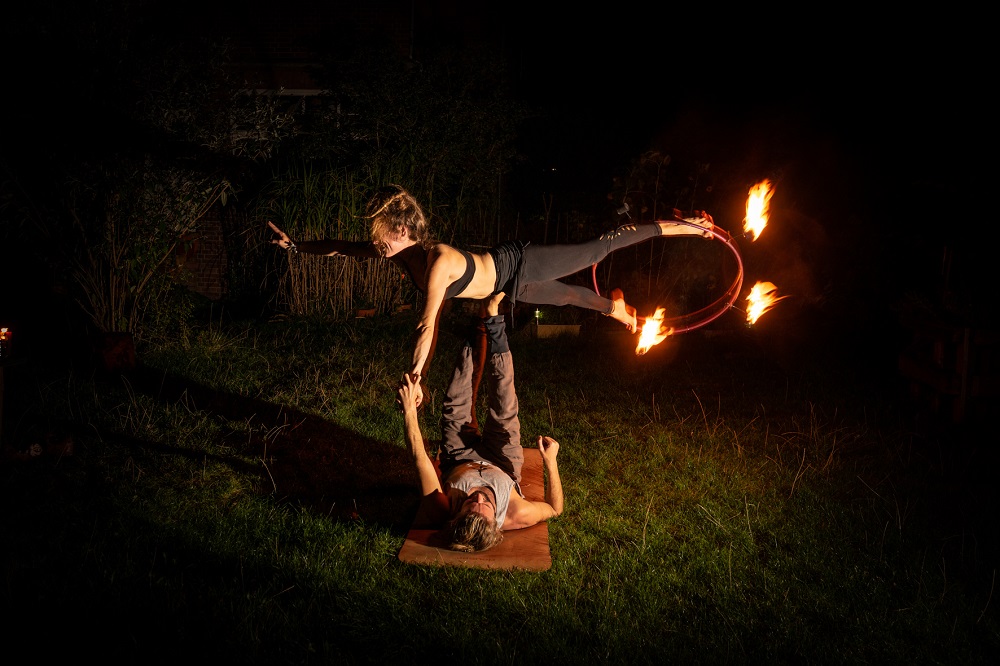 AcroYoga-Side-Star: Eine Fliegerin balanciert seitlich auf den Füßen ihres Partners, ein brennender Feuerreifen glüht an ihren Füßen