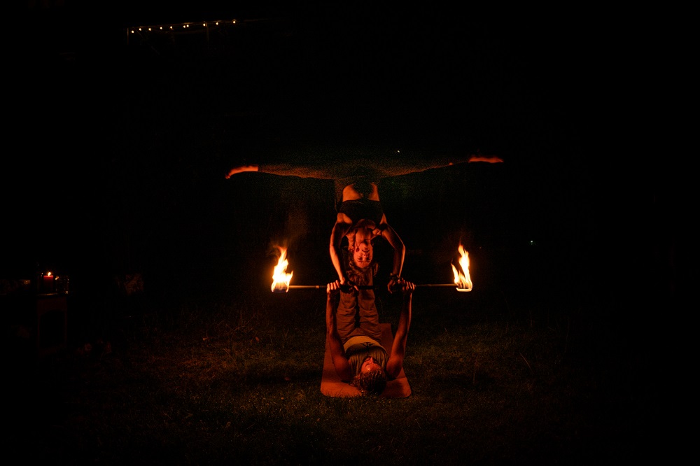 AcroYoga-Star-Pose: Eine Fliegerin balanciert waagerecht auf den Füßen ihres Partners, zwischen beiden ein Feuerstab