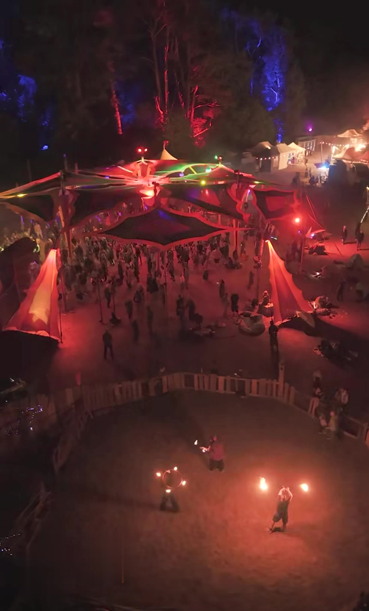 Luftaufnahme des Firespace auf dem New Healing Festival bei Nacht: Feuerkünstler in einem eingezäunten Feuerkreis, dahinter eine große Tanzfläche unter Sonnensegeln mit Festivalgästen